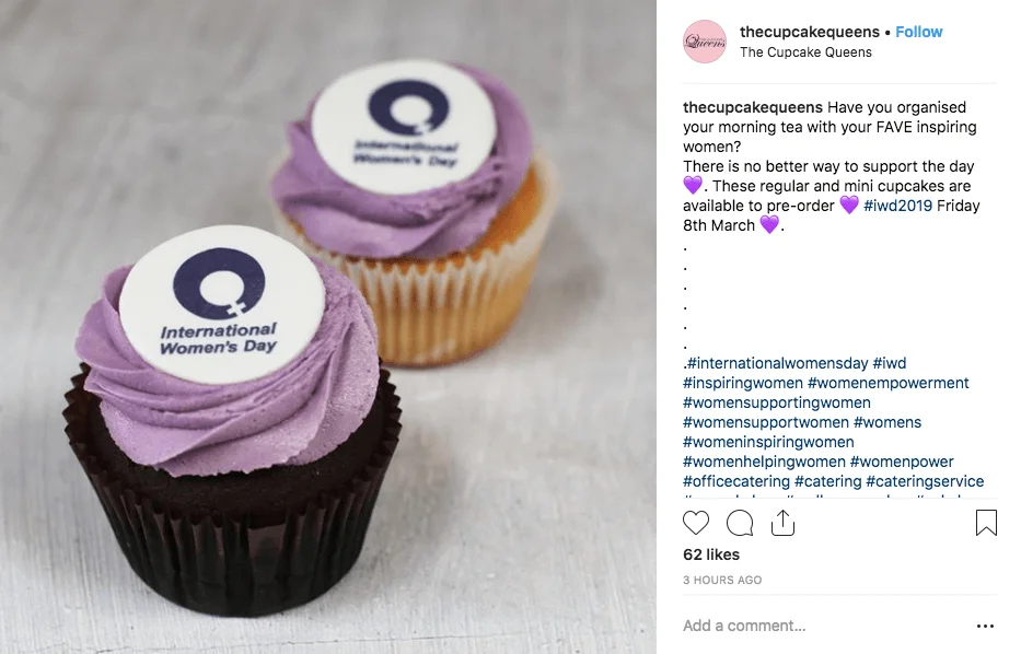 Instagram post showing two cupcakes decorated with purple icing and the International Women's Day logo. The cakes are available to pre-order for IWD celebrations.