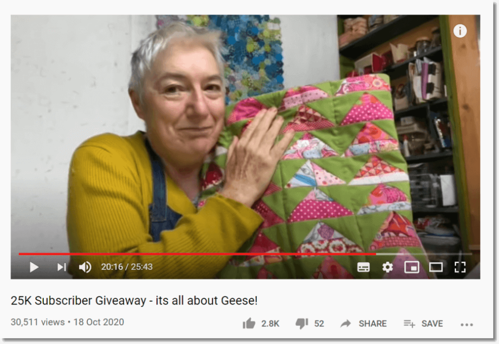 Screenshot of a YouTube video titled "25K subscriber giveaway - it's all about geese!" The picture shows a woman with short white hair in a yellow jumper, smiling and holding up a pink and green quilted cushion cover.